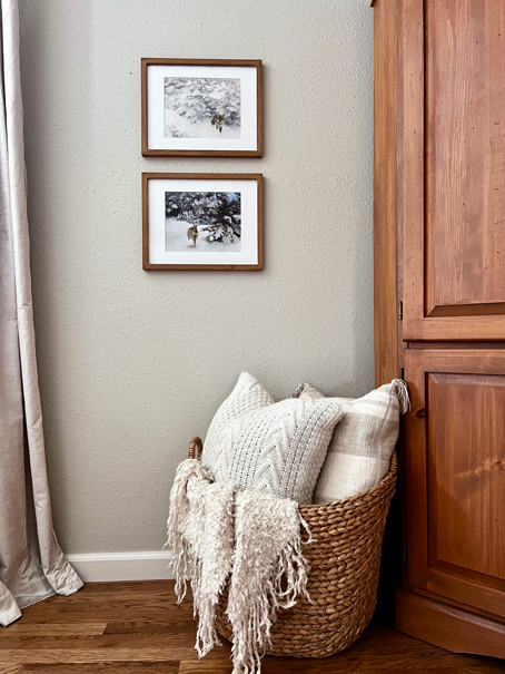 A basket sits on the floor filled with varying textures of neutral pillows and blankets. Above is two winter paintings of a fox and a rabbit. 