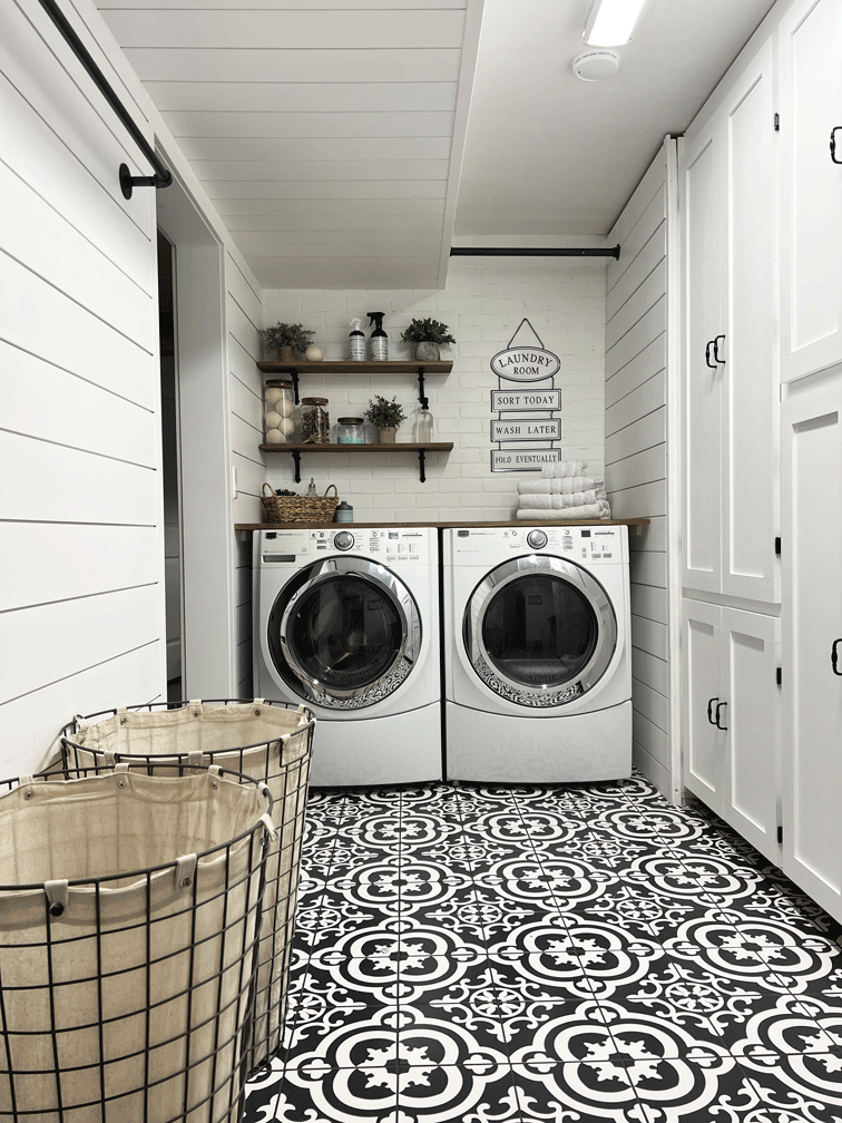 Functional laundry room ideas for a laundry room with built in storage, bar height folding table, drying racks for hanging or hang drying laundry, dirty laundry metal storage baskets, and decorated with woven baskets, a laundry room sign, a woven rug, and patterned tile to compliment the wood and white board and batten and shiplap walls.