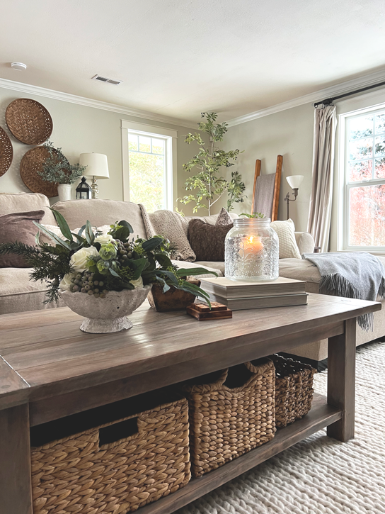 Neutral living room scene with a coffee table and a winter bouquet of pine and white roses and a candle glowing from within a jar. On the couch lies cozy knit blankets, faux fur pillow covers and a knit pillows. A olive tree sits in the background with a blanket ladder. 