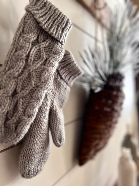 Cozy Hygge-Inspired Winter Living -A pair of cozy knit mittens sits in front of a woven hanging basket and pine stems. 