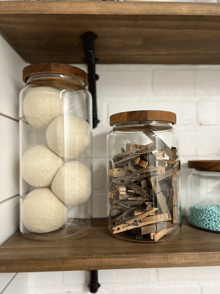 Glass canisters are filled with wool dryer balls, wood clothes pins, and laundry beads. Plants and soap dispensters/ linen sprays in pretty containers also sit atop wood shelving with black iron brackets against a white brick wall. 