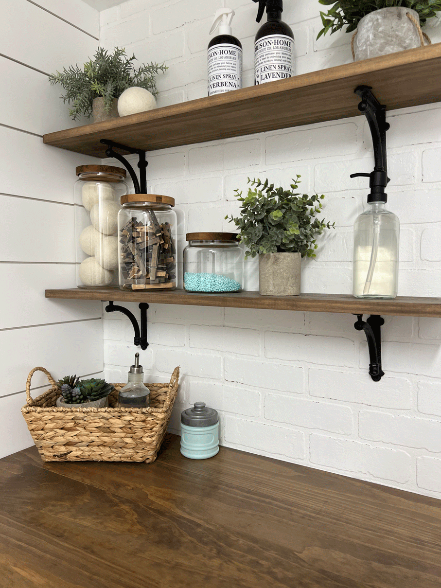 Open shelving. Glass canisters are filled with wool dryer balls, wood clothes pins, and laundry beads. Plants and soap dispensters/ linen sprays in pretty containers also sit atop wood shelving with black iron brackets against a white brick wall. 