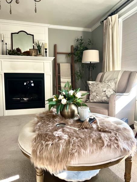 A Cozy Hygge-Inspired Winter Living view of a cup of hot tea, a magnifying glass, a candle in a votive, and a bouquet or roses and eucalyptus all resting atop a faux fur rug. A side chair sits along side with a mantel and mirror in the background. A blanket ladder, floor lamp and olive tree sit in the corner. 