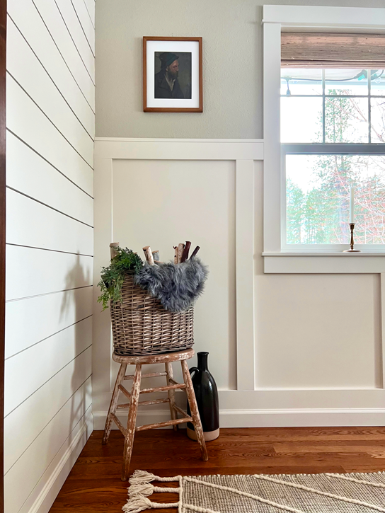 Shiplap and board and batten walls sit as a backdrop to a chippy painted antique stool with a basket of fuax fur, pine, and wood sticks. A vintage portrait of a man is on the wall above with a vintage jug on the floor beside. 
