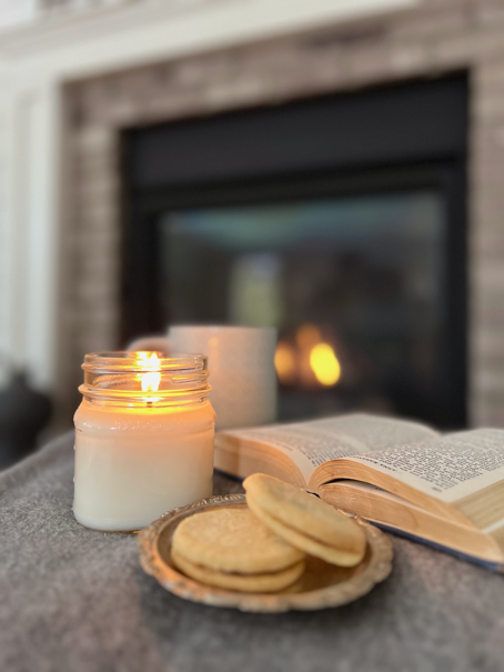 A lit candle jar sits next to an open book, a cup of hot coffee and a few butter cookies while a fire glows in the background. 