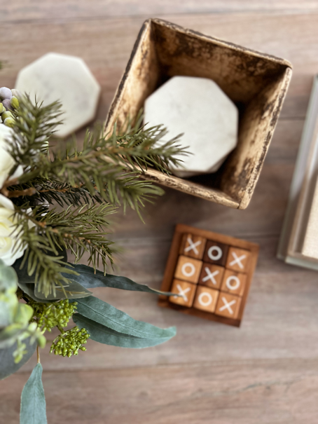 Cozy Hygge-Inspired Winter Living -A flatlay view of a cup of tea, an open book, a pine and white rose bouquet, a lit candle glowing from within an embossed jar, and a wooden tic tac toe game. 