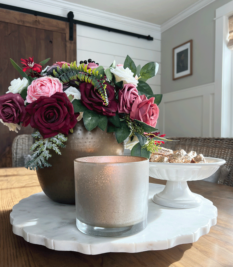 DIY Mercury Glass Votives: A small candle holder in shimmery gold, silver, metallic pink, and copper colors. Candles glow from within, and a rich-colored burgundy and pink bouquet in a gold vase sit in the background. 